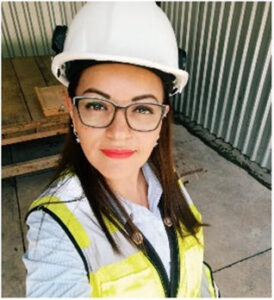 Smiling woman wearing a white hard hat, glasses, and a yellow safety vest at a construction site