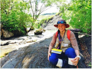 Smiling woman wearing a hat and outdoor gear, sitting on a rock by a stream surrounded by lush greenery