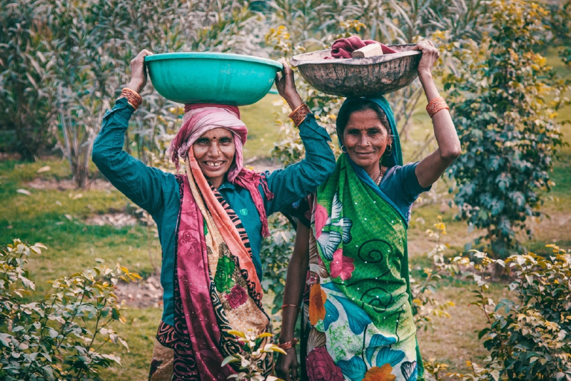 Two smiling women in colorful traditional attire carrying baskets on their heads in a lush garden setting